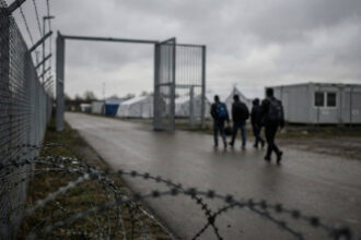 Εξωτερική άποψη κέντρου υποδοχής και καταγραφής αιτούντων άσυλο στη Γερμανία, με συρματοπλέγματα και θολές φιγούρες στο βάθος.