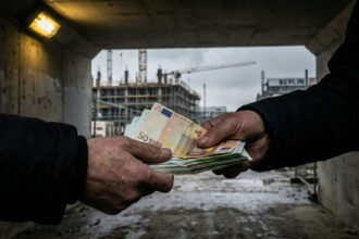 Συμβολική απεικόνιση της παραοικονομίας με σκιώδεις φιγούρες να ανταλλάσσουν χαρτονομίσματα ευρώ μπροστά από ένα θολό εργοτάξιο στο Βερολίνο.