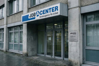 Κοντινό πλάνο στην πρόσοψη ενός γερμανικού κέντρου εργασίας (Jobcenter) με το λογότυπο να φαίνεται καθαρά, υπό συννεφιασμένο ουρανό.