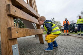 Παιδί με κίτρινο γιλέκο ασφαλείας και αδιάβροχο παντελόνι επιθεωρεί ξύλινη παιδική χαρά
