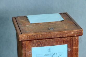 Κοντινό πλάνο σε χέρια που κρατούν εκλογικό φάκελο με την ελληνική σημαία στο φόντο, θολή εικόνα του Υπουργείου Εσωτερικών.
