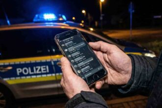 Smartphone με ανοιχτή εφαρμογή κοινωνικής δικτύωσης και φόντο αστυνομικά φώτα στη Γερμανία