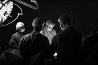 Δύο χειρουργοί με ιατρικές μάσκες και ειδικές στολές στέκονται σιωπηλοί μέσα σε έναν σκοτεινό θάλαμο χειρουργείου