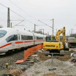 Ένα σύγχρονο λευκό τρένο ICE σταματημένο σε ράγες δίπλα σε εργοτάξιο με ένα κίτρινο μηχάνημα εκσκαφής