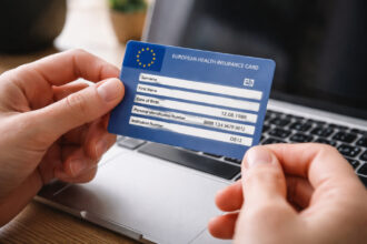 Eine Person tippt auf einem Laptop und hält eine blaue Europäische Krankenversicherungskarte in der Hand.
