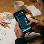 Eine Person hält ein Smartphone mit einer Online-Casino-App in der Hand, im Hintergrund sind unscharf Euro-Banknoten und Steuerdokumente zu sehen.