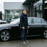 Ένας άνδρας στέκεται δίπλα σε μια μαύρη πολυτελή Mercedes έξω από μια σύγχρονη αντιπροσωπεία αυτοκινήτων