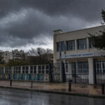 Dunkle Gewitterwolken über einer griechischen Stadt, starker Regen fällt auf eine leere Straße vor einem geschlossenen Schulgebäude.