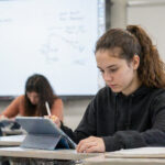 Ein Schüler nutzt ein Tablet an einem modernen Schulplatz, im Hintergrund ist eine interaktive digitale Tafel zu sehen.