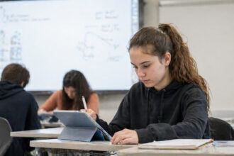 Ein Schüler nutzt ein Tablet an einem modernen Schulplatz, im Hintergrund ist eine interaktive digitale Tafel zu sehen.