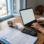 Eine Person füllt eine Steuererklärung am Computer aus, daneben liegen Dokumente der AADE