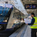 Ein moderner Hochgeschwindigkeitszug steht in einem griechischen Bahnhof, während Techniker in Warnwesten auf dem Bahnsteig Pläne besprechen.