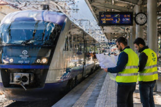 Ein moderner Hochgeschwindigkeitszug steht in einem griechischen Bahnhof, während Techniker in Warnwesten auf dem Bahnsteig Pläne besprechen.
