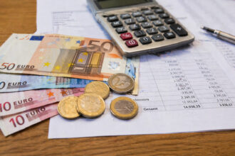 Euroscheine und Münzen liegen auf einem Tisch neben Formularen und einem Taschenrechner