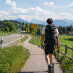 Ein junger Mann mit einem großen Rucksack wandert auf einem Radweg entlang einer bayerischen Landstraße.