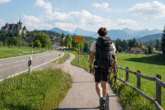 Ein junger Mann mit einem großen Rucksack wandert auf einem Radweg entlang einer bayerischen Landstraße.