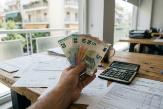 Eine Hand hält Euro-Banknoten vor dem Hintergrund griechischer Arbeitsverträge und Finanzdokumente.