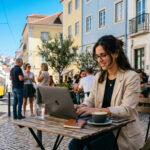 Ein junger Berufstätiger arbeitet konzentriert an seinem Laptop auf einer sonnigen Terrasse in Griechenland.