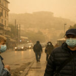 Eine dichte, gelbliche Wolke aus Saharastaub hängt über einer städtischen Straße in Griechenland, während Fußgänger Schutzmasken tragen.