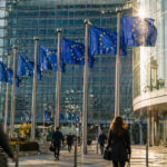 Finanzzentrum mit europäischen Flaggen und einer Anzeigetafel für Aktienkurse an einem sonnigen Tag.