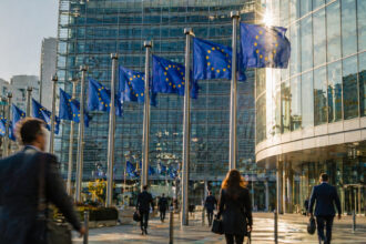 Finanzzentrum mit europäischen Flaggen und einer Anzeigetafel für Aktienkurse an einem sonnigen Tag.