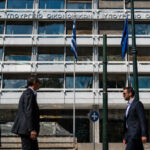 Außenansicht des griechischen Finanzministeriums in Athen an einem sonnigen Tag mit Geschäftsleuten im Vordergrund.