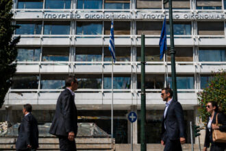 Außenansicht des griechischen Finanzministeriums in Athen an einem sonnigen Tag mit Geschäftsleuten im Vordergrund.