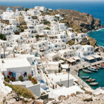 Eine dicht bebaute Küstenlinie einer Kykladeninsel mit weißen Häusern, die den Hang hinunter bis zum Meer reichen.