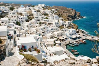 Eine dicht bebaute Küstenlinie einer Kykladeninsel mit weißen Häusern, die den Hang hinunter bis zum Meer reichen.