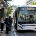 Ein moderner elektrischer Stadtbus fährt durch eine belebte Straße in Athen, Fahrgäste warten an einer modernen Haltestelle.