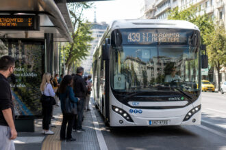 Ein moderner elektrischer Stadtbus fährt durch eine belebte Straße in Athen, Fahrgäste warten an einer modernen Haltestelle.
