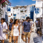 Eine Gruppe von Touristen spaziert bei sonnigem Wetter durch eine malerische griechische Gasse mit weißen Häusern.