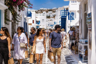 Eine Gruppe von Touristen spaziert bei sonnigem Wetter durch eine malerische griechische Gasse mit weißen Häusern.