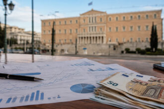 Finanzdiagramme und Euroscheine auf einem Tisch mit Blick auf ein griechisches Regierungsgebäude