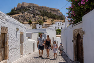Eine Familie spaziert durch die autofreien, gepflasterten Gassen von Lindos auf Rhodos, im Hintergrund weiße Häuser.