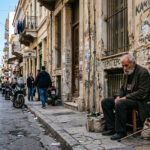 Ein älterer Mann sitzt auf dem Gehweg in einer Straße in Athen, was die Problematik der Obdachlosigkeit verdeutlicht.
