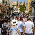 Viele Touristen spazieren an einem sonnigen Tag durch eine belebte Straße im historischen Zentrum von Athen, während im Hintergrund ein Bus vorbeifährt.