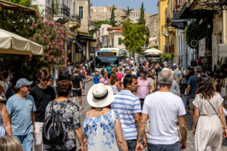Viele Touristen spazieren an einem sonnigen Tag durch eine belebte Straße im historischen Zentrum von Athen, während im Hintergrund ein Bus vorbeifährt.