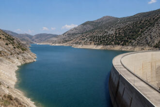 Ein gefüllter Stausee in Griechenland, umgeben von trockener mediterraner Hügellandschaft unter strahlend blauem Himmel.