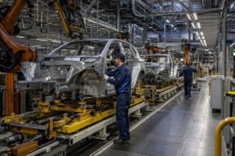 Σύγχρονο γερμανικό εργοστάσιο αυτοκινήτων με ρομποτικούς βραχίονες και εργάτες στη γραμμή παραγωγής.