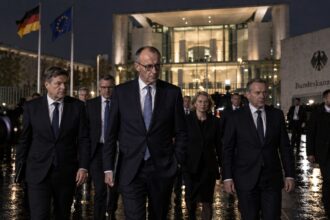 Πολιτικοί με κοστούμια συνομιλούν έξω από κυβερνητικό κτίριο στο Βερολίνο τη νύχτα.