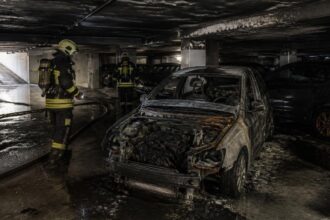 Καμένο αυτοκίνητο σε υπόγειο πάρκινγκ με πυροσβέστες να εξετάζουν τον χώρο.