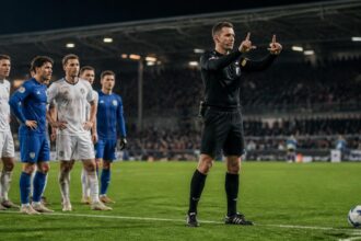 Διαιτητής κάνει το σήμα του VAR δίπλα σε ποδοσφαιριστές που περιμένουν την απόφασή του σε γήπεδο.