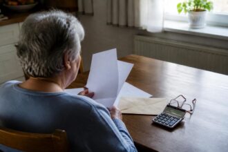 Άτομο καθισμένο σε τραπέζι σπιτιού εξετάζει έγγραφα δίπλα σε φάκελο και αριθμομηχανή