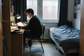 Φοιτητής μελετά σε μικρό δωμάτιο συγκατοίκησης με γραφείο, βιβλία και κρεβάτι