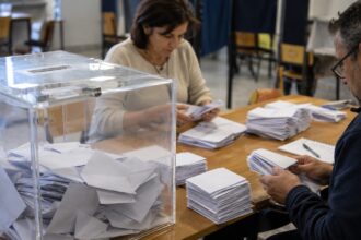 Δύο άνθρωποι ταξινομούν διπλωμένα ψηφοδέλτια σε μακρύ τραπέζι δίπλα σε διάφανη κάλπη.