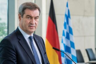 Συμβολικό πορτρέτο του Markus Söder σε γερμανικό πολιτικό περιβάλλον με σημαίες στο βάθος