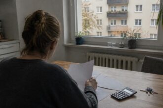 Κάτοικος εξετάζει έγγραφα στεγαστικών εξόδων δίπλα σε αριθμομηχανή μέσα σε διαμέρισμα